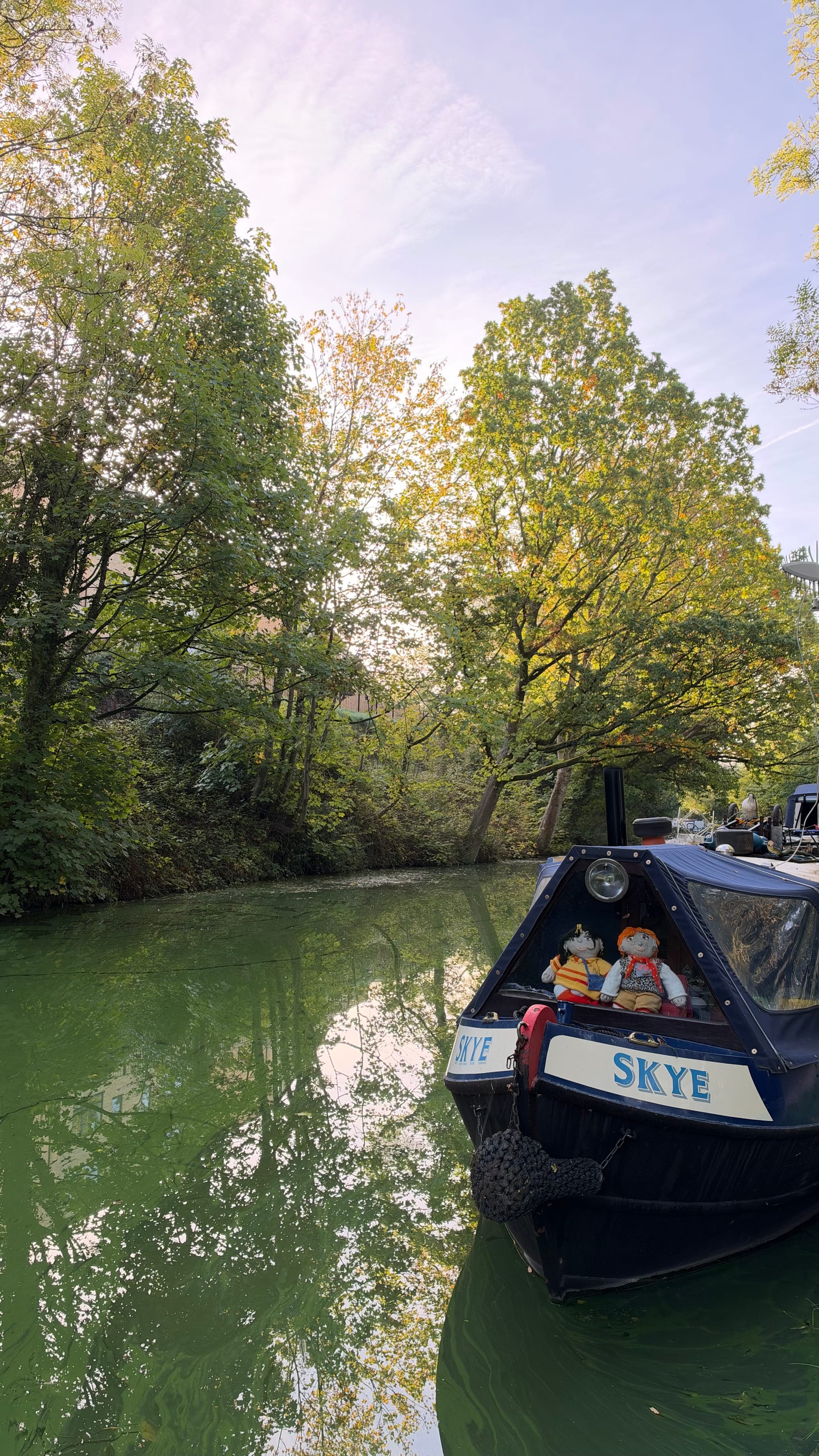 A still stretch of the Leeds and Liverpool Canal on an early autumn morning. Tall trees, mostly green with the first yellow and orange leaves, line the far bank and are mirrored in the calm green water. A pale lilac sky shows through the branches. In the foreground on the right is the bow of a navy blue narrowboat named Skye, with two knitted dolls sitting in the front window, one with dark hair and one with ginger hair.
