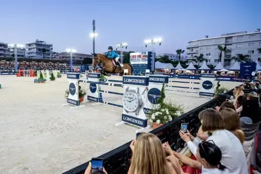 Binomio saltando un obstáculo con branding Longines durante el Gran Premio de Cannes, con público fotografiando desde la tribuna.