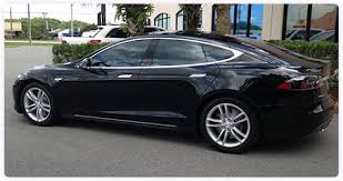 Side view of a black Tesla Model S parked outdoors near palm trees and a glass-fronted building.