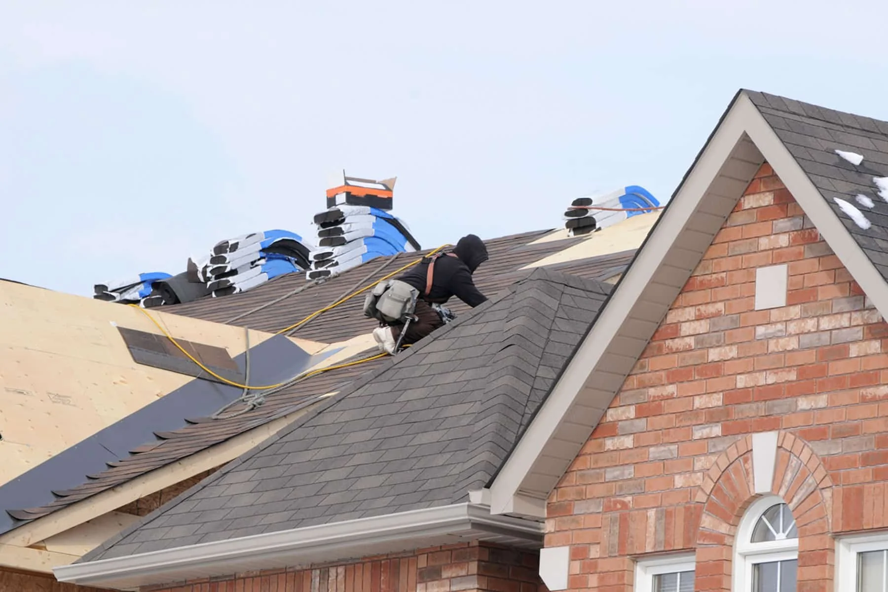 Worker installing roof shingles on a snowy residential roof using safety harness and gear.