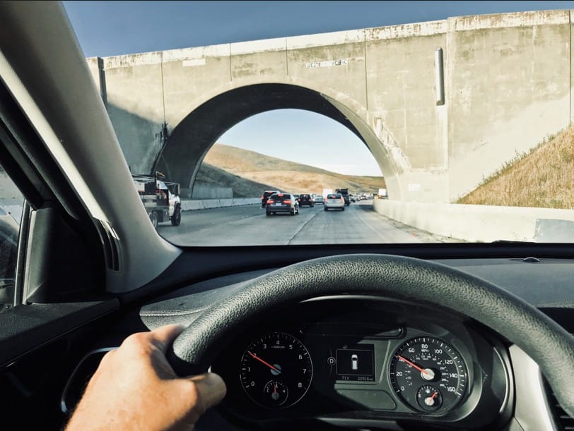 A first-person view from the driver's seat of a car traveling on a busy highway. The vehicle is approaching a large concrete arched overpass with "Western Pacific" inscribed on it. Other cars are visible in the lanes ahead under a clear blue sky.