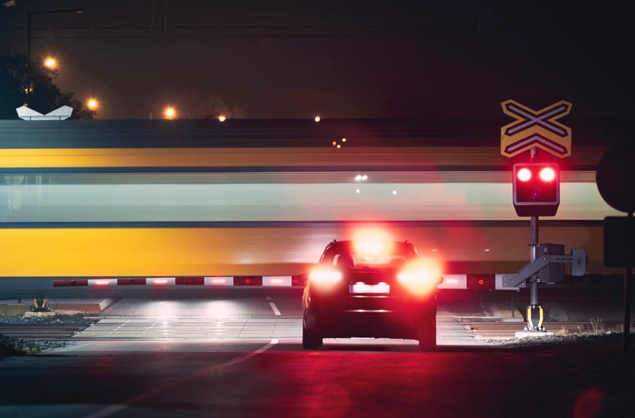 A car waits at a closed railroad crossing at night as a yellow and grey train speeds past, blurred by motion. Red warning lights are illuminated on the crossing signal.