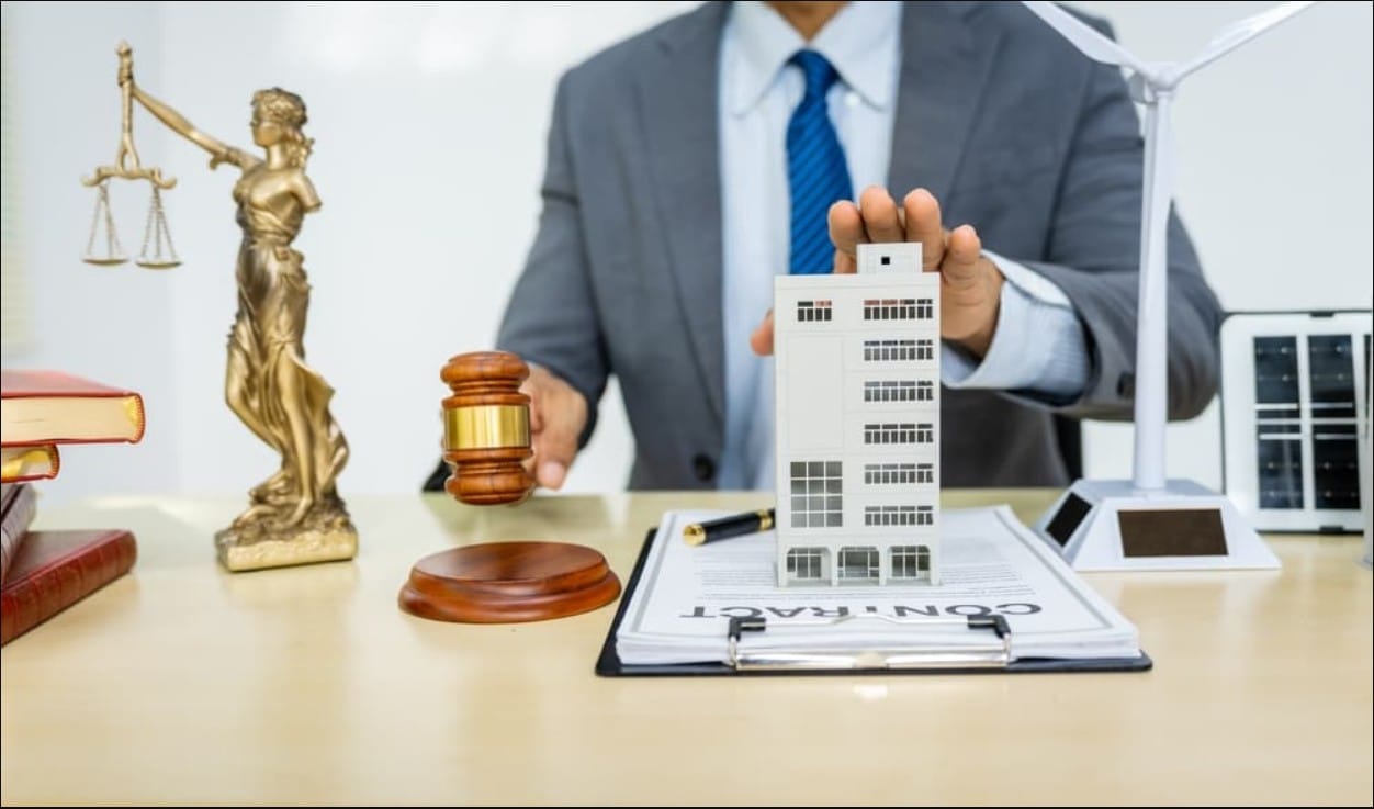 A lawyer in a suit holding a wooden gavel next to a model building, a contract, and a Lady Justice statue, representing real estate or environmental law.