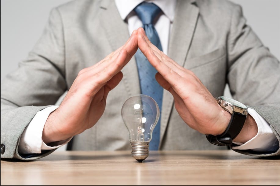 A person in a grey business suit forming a protective roof shape with their hands over a lightbulb on a wooden desk.