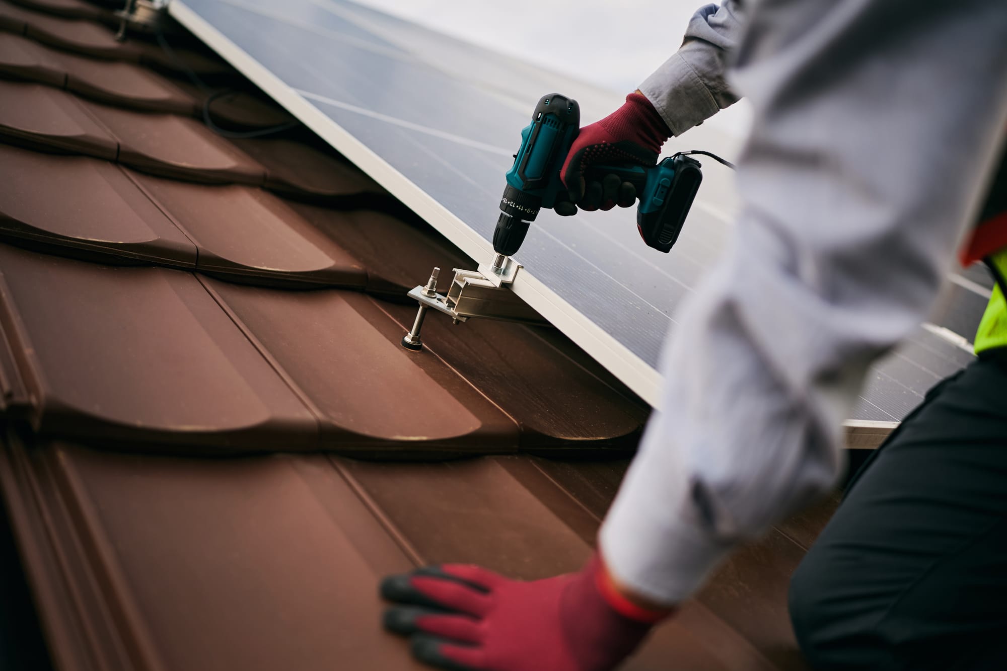 A technician using a power drill to install solar panels onto a brown tile roof.