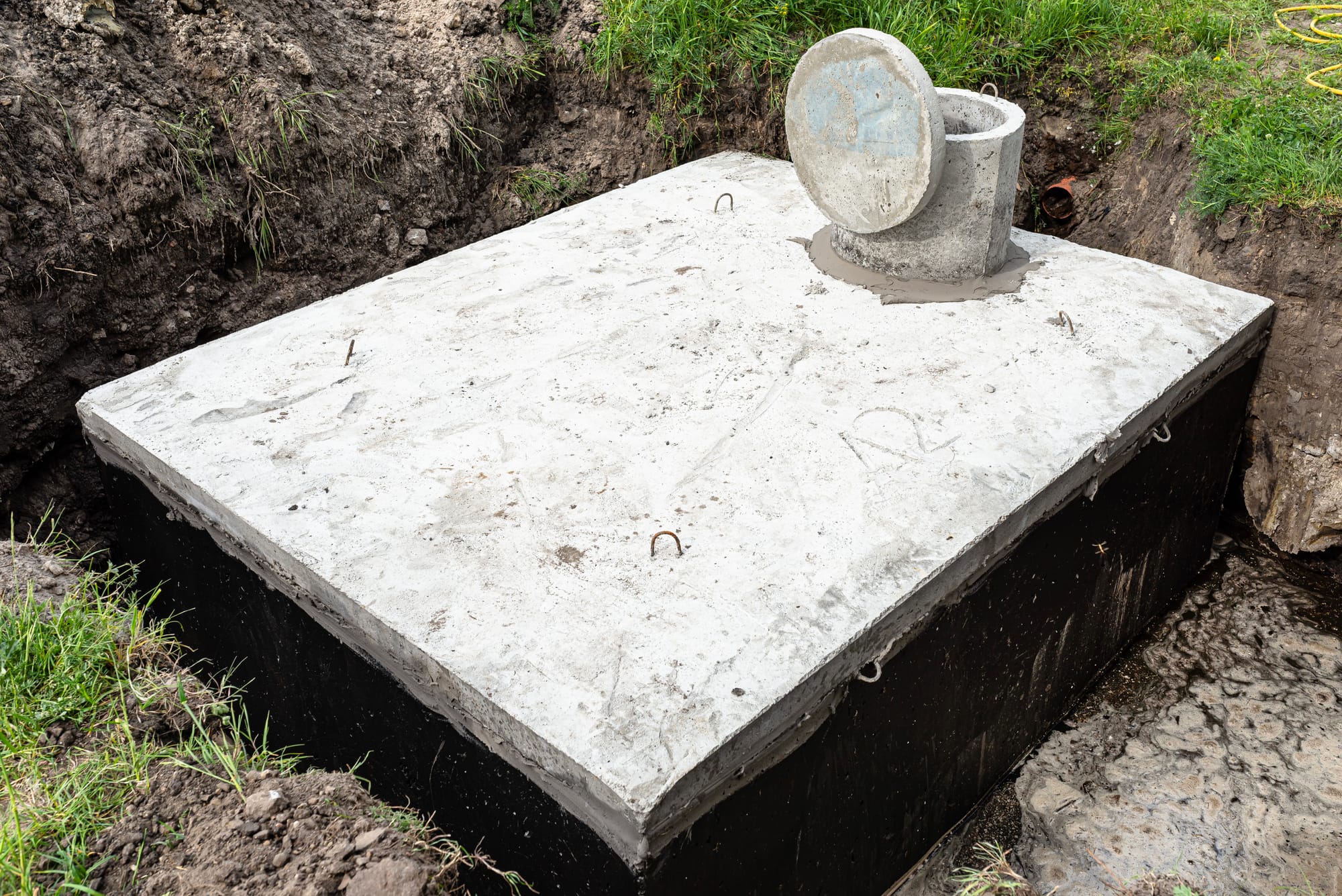 A rectangular concrete septic tank installed in a dirt excavation, featuring a round concrete riser with an open lid on top.
