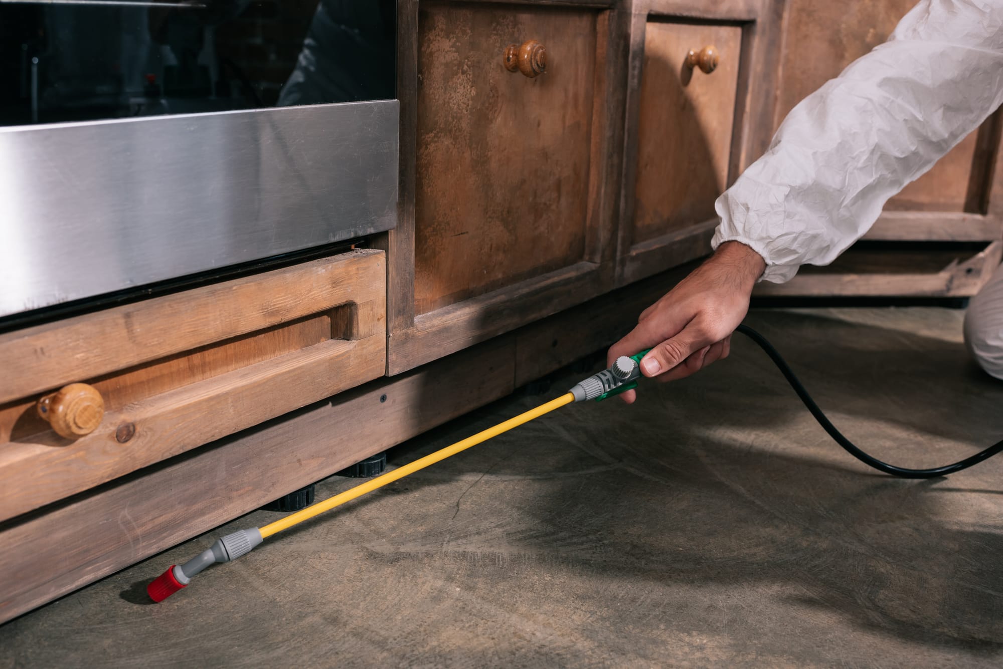 A person in a white protective suit using a long yellow spray wand to apply pest control treatment along the baseboards of wooden kitchen cabinets.