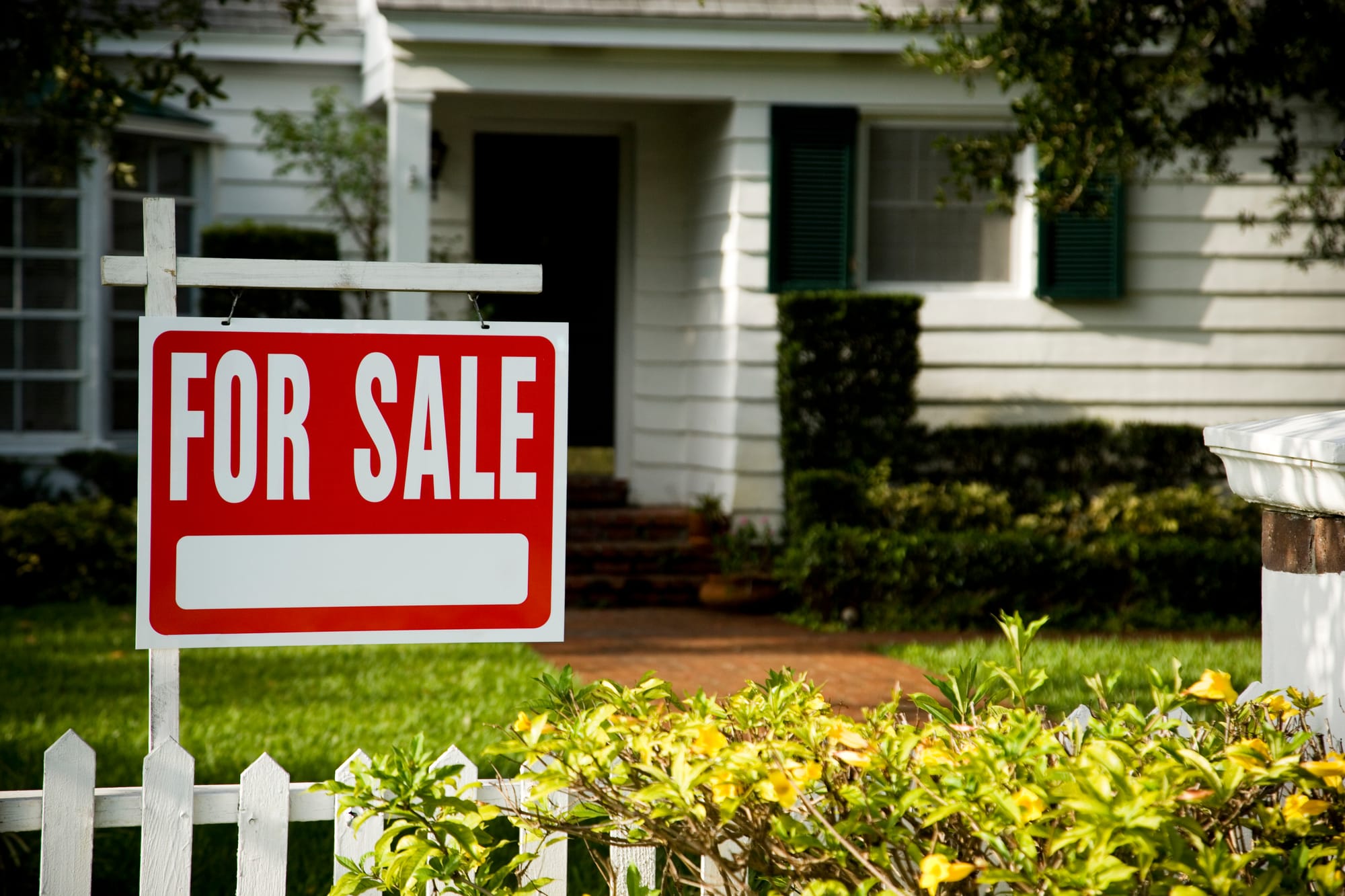 A red and white “For Sale” sign displayed in the front yard of a suburban home with a walkway leading to the entrance.