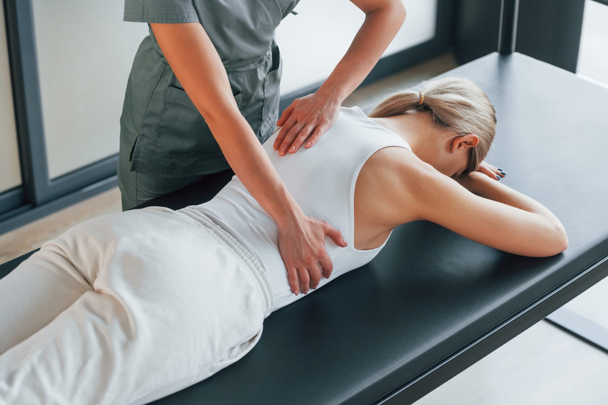 A physical therapist in grey scrubs performing a manual back adjustment on a woman lying face down on a professional treatment table.