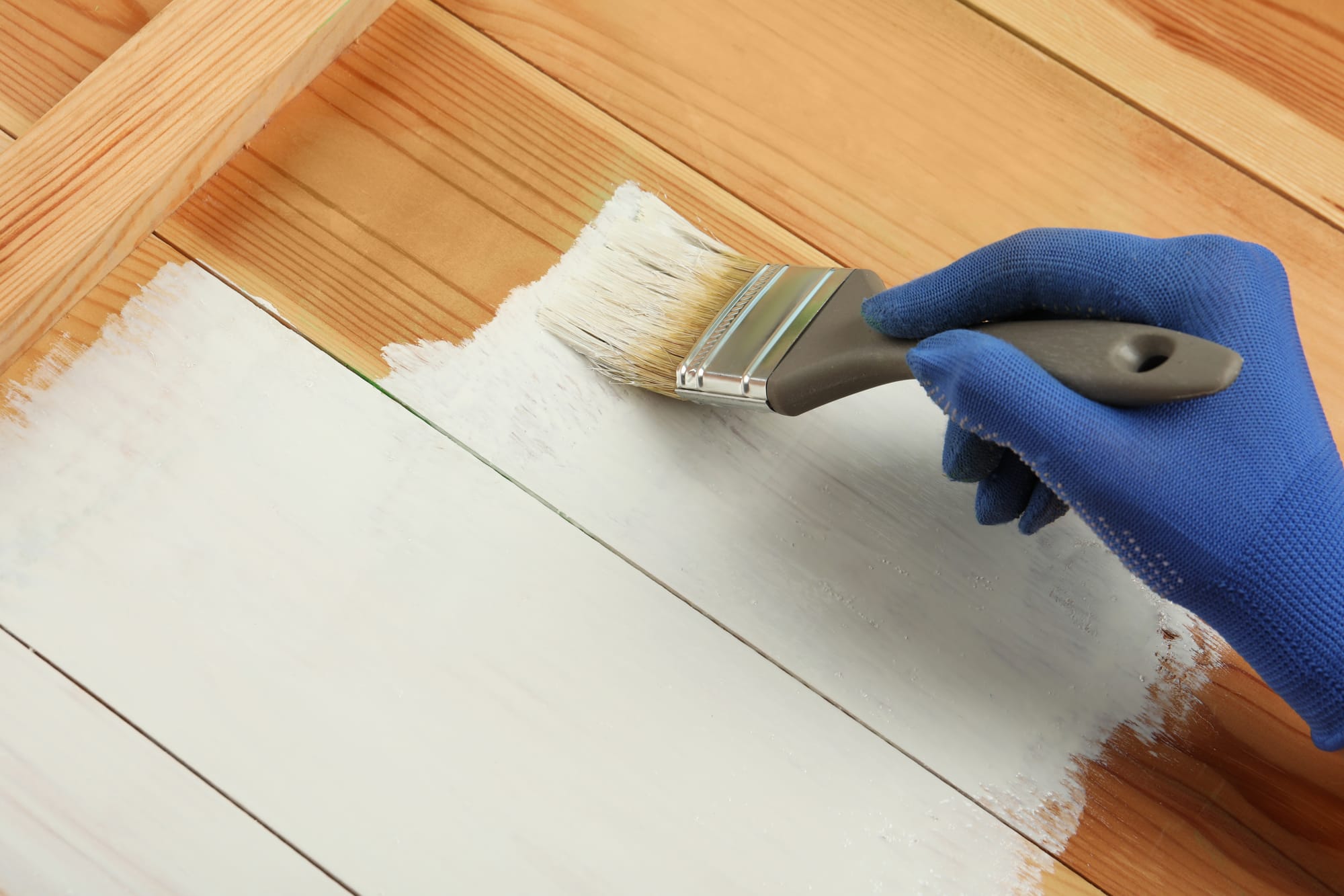 hand in a blue glove using a paintbrush to apply white paint to wooden planks.