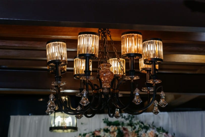 Elegant black metal chandelier with warm glowing crystal cylinders and hanging glass droplets, set against a dark wooden beamed ceiling.