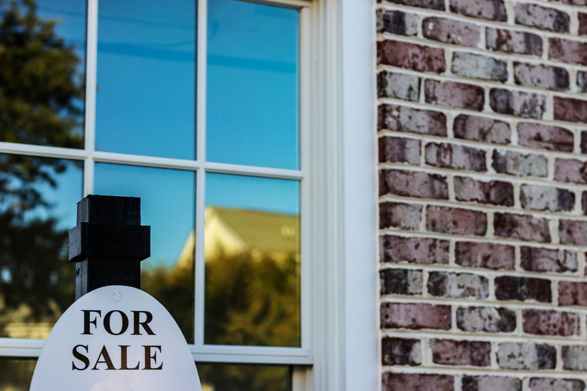 A “For Sale” sign stands in front of a brick house with a large window, suggesting a residential property on the market.