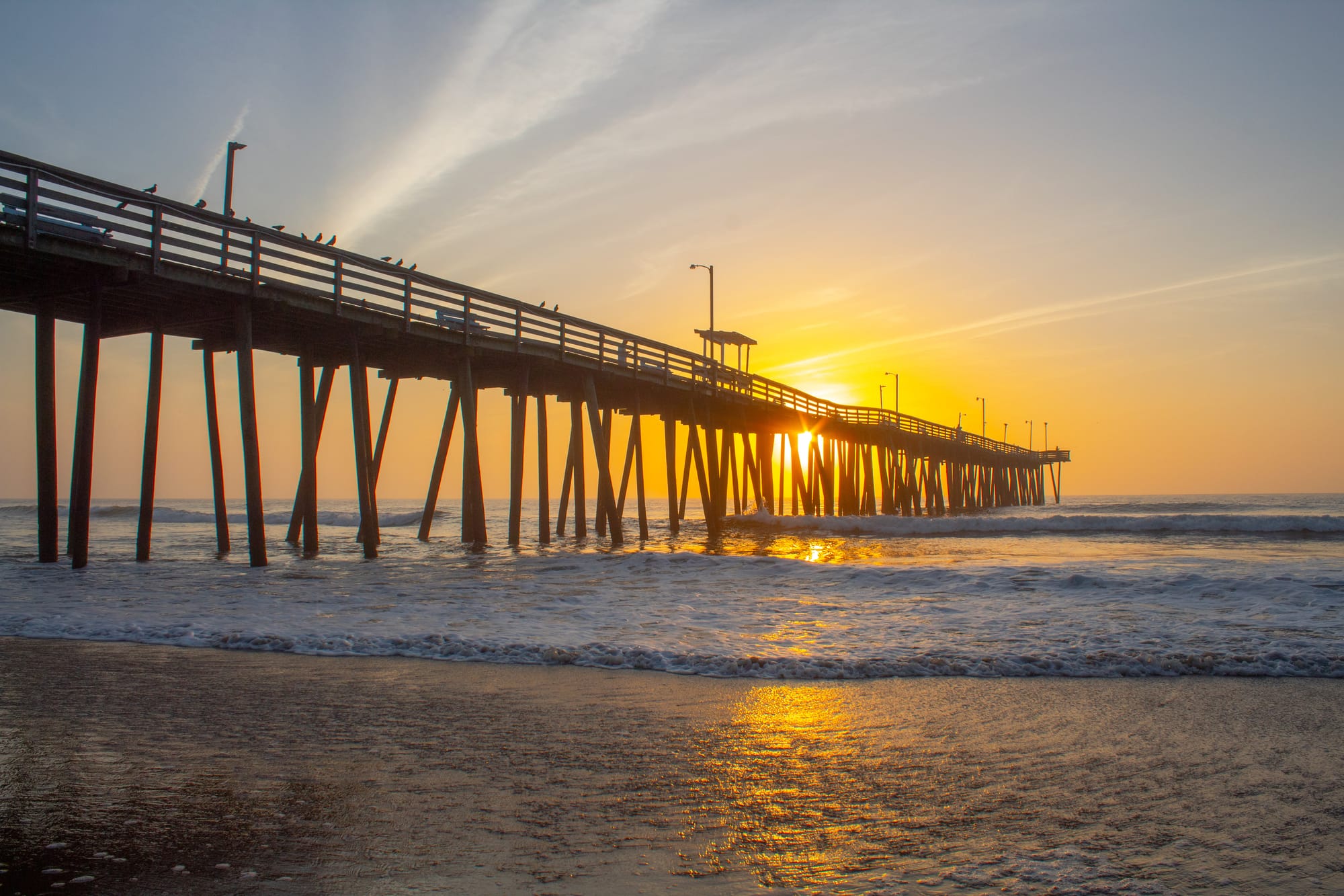 Sunrise over the ocean beneath a long wooden pier in Virginia Beach, with waves rolling onto the shore and warm golden light reflecting on the water.