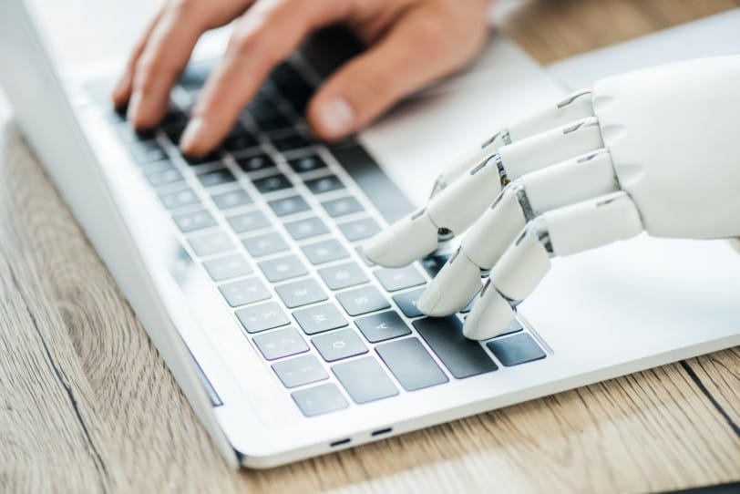 A close-up view of a human hand and a white robotic hand side by side typing on a laptop keyboard, symbolizing human-AI collaboration.