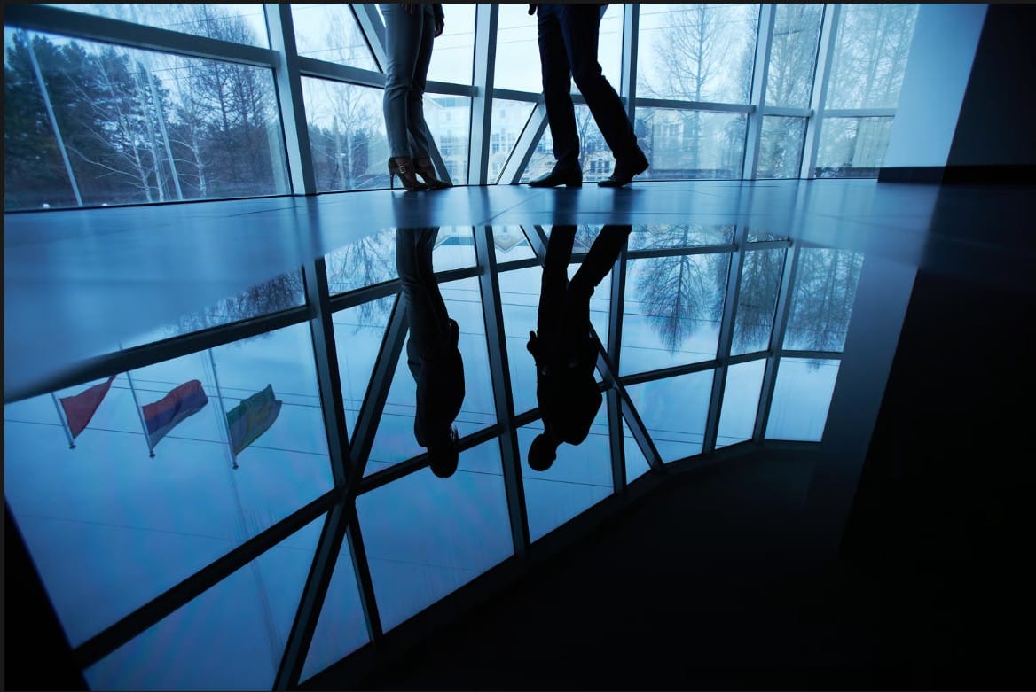 Low-angle reflection of two corporate colleagues standing together in a modern glass office building corridor.