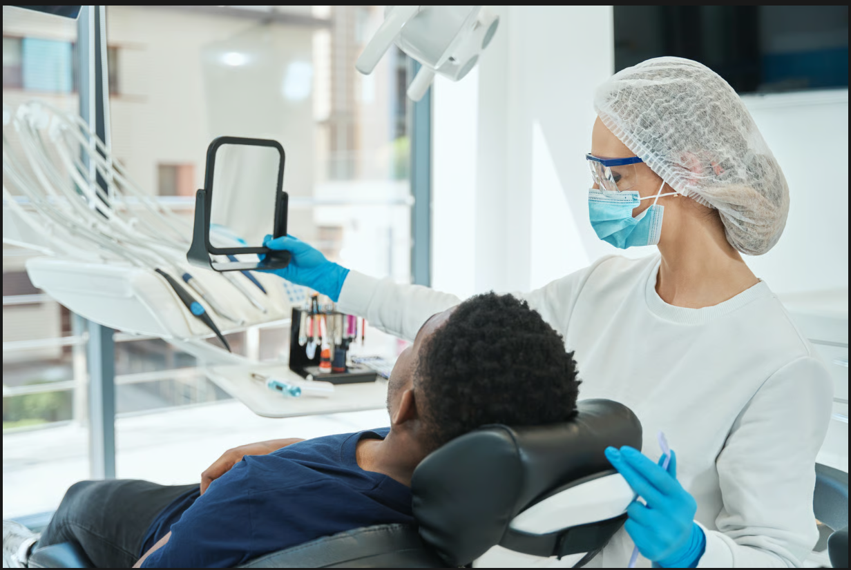 Dentist wearing protective gear holding a handheld mirror for a patient reclining in the exam chair.