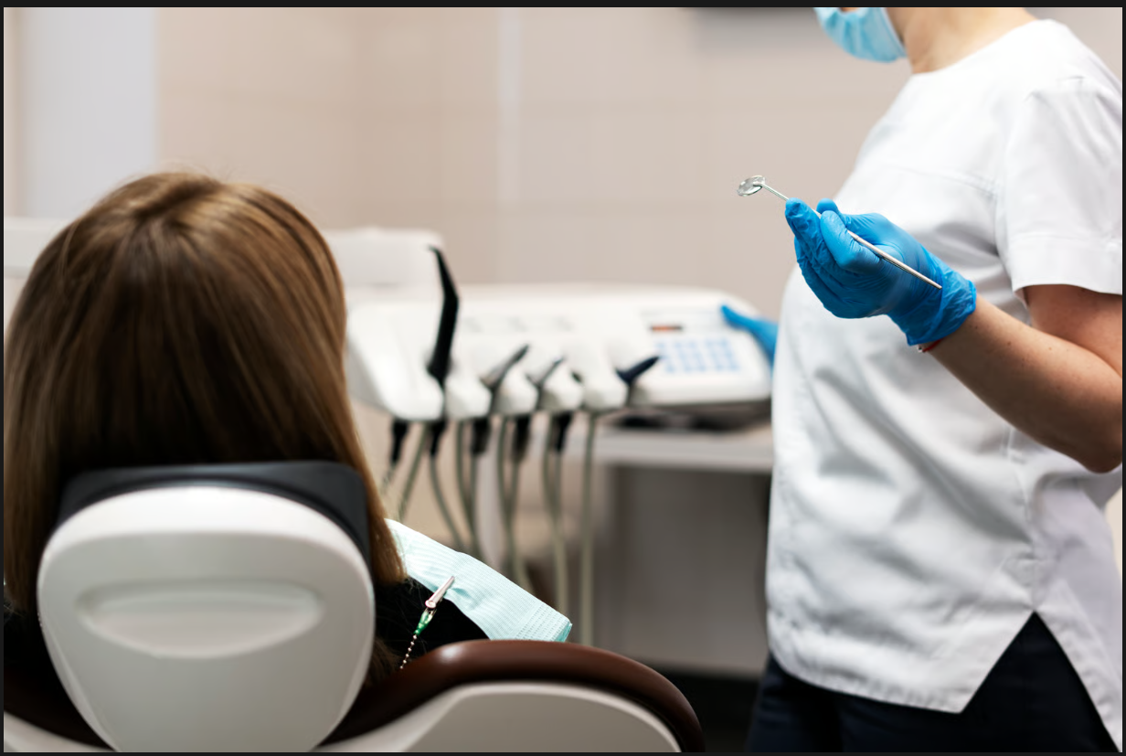 Dentist in blue gloves and white uniform holding a mouth mirror next to a patient in the dental chair.