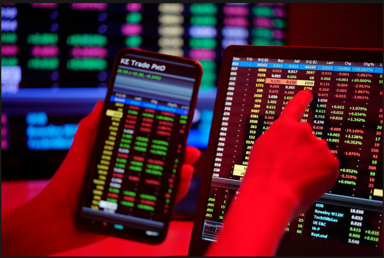 Trader holding a smartphone and pointing at a tablet displaying real-time stock market data and trade figures.