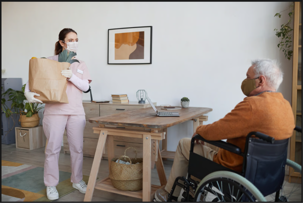 A caregiver wearing a mask, scrubs, and gloves delivers a large paper bag of fresh vegetables to an elderly senior man in a wheelchair, illustrating safe home care practices and health safety protocols.