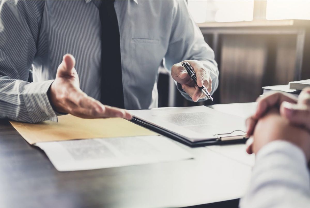 A professional in a shirt and tie gestures toward legal documents and a contract on a desk while holding a pen during a consultation.