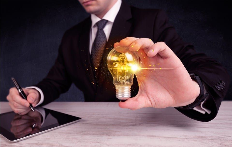 A person in a business suit holding a glowing, lit-up lightbulb while sitting at a desk with a tablet and pen.