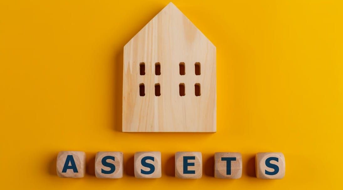 A wooden house cutout placed above wooden blocks spelling the word "ASSETS" against a solid yellow background.