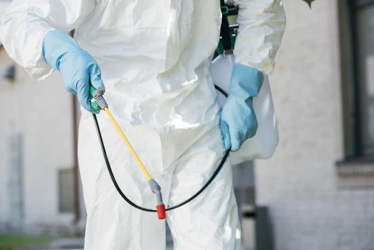 A person in a white protective hazmat suit and blue gloves holding a yellow spray wand attached to a pressurized tank for outdoor pest control.