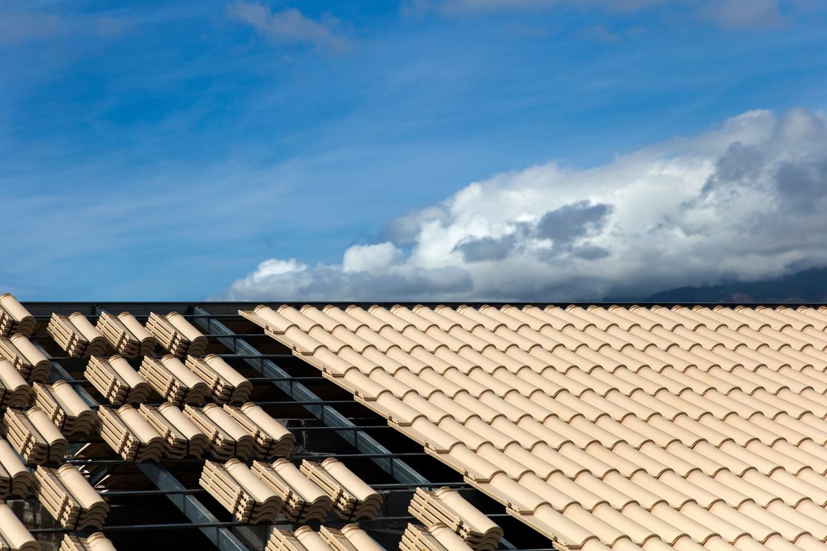 Tan clay roof tiles being installed on a metal structural frame against a blue sky with white clouds.