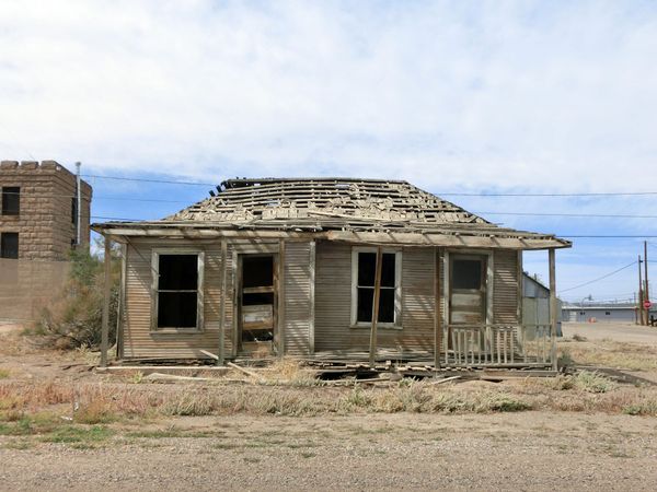 Old abandoned wooden house with collapsed roof, broken porch, and missing windows, set in a rural town with utility poles and sparse vegetation.