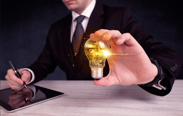 A person in a business suit holding a glowing, lit-up lightbulb while sitting at a desk with a tablet and pen.