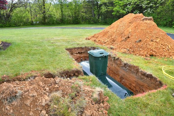 A large green septic tank being installed in a deep rectangular trench in a grassy yard, next to a large pile of excavated orange soil.
