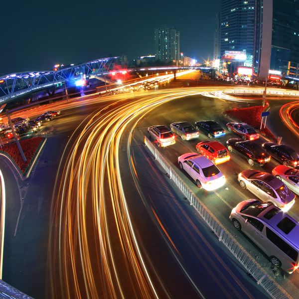 High-angle long-exposure photo of a city intersection at night. Orange and white light trails curve through traffic, with stationary cars and glowing skyscrapers in the background.