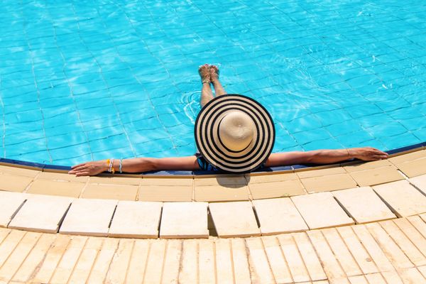 An overhead view of a person wearing a large black-and-white striped sun hat, relaxing at the edge of a bright blue tiled swimming pool with their arms outstretched along the stone coping.