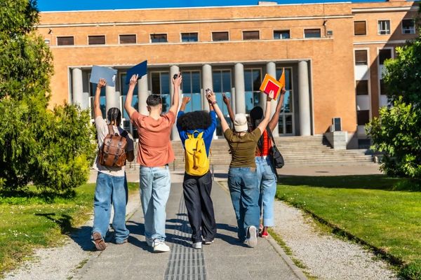 Diverse group of college students holding up notebooks and cheering while walking toward a campus building.