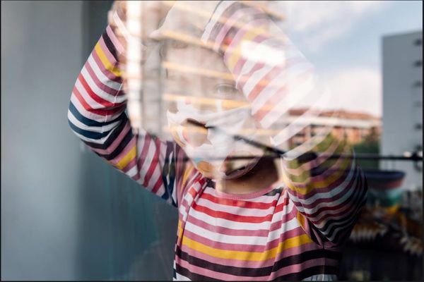 Child in a colorful striped long-sleeve shirt and face mask with hands raised near a window reflection.