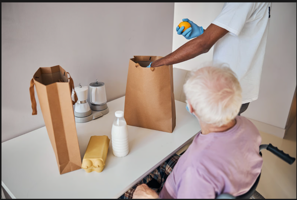 Masked home care assistant wearing protective gloves unpacking fresh groceries for a senior homeowner following strict health safety protocols.