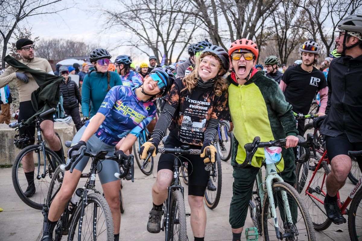Three people in bike helmets pose with enthusiastic facial expressions in front of a crowd of cyclists.