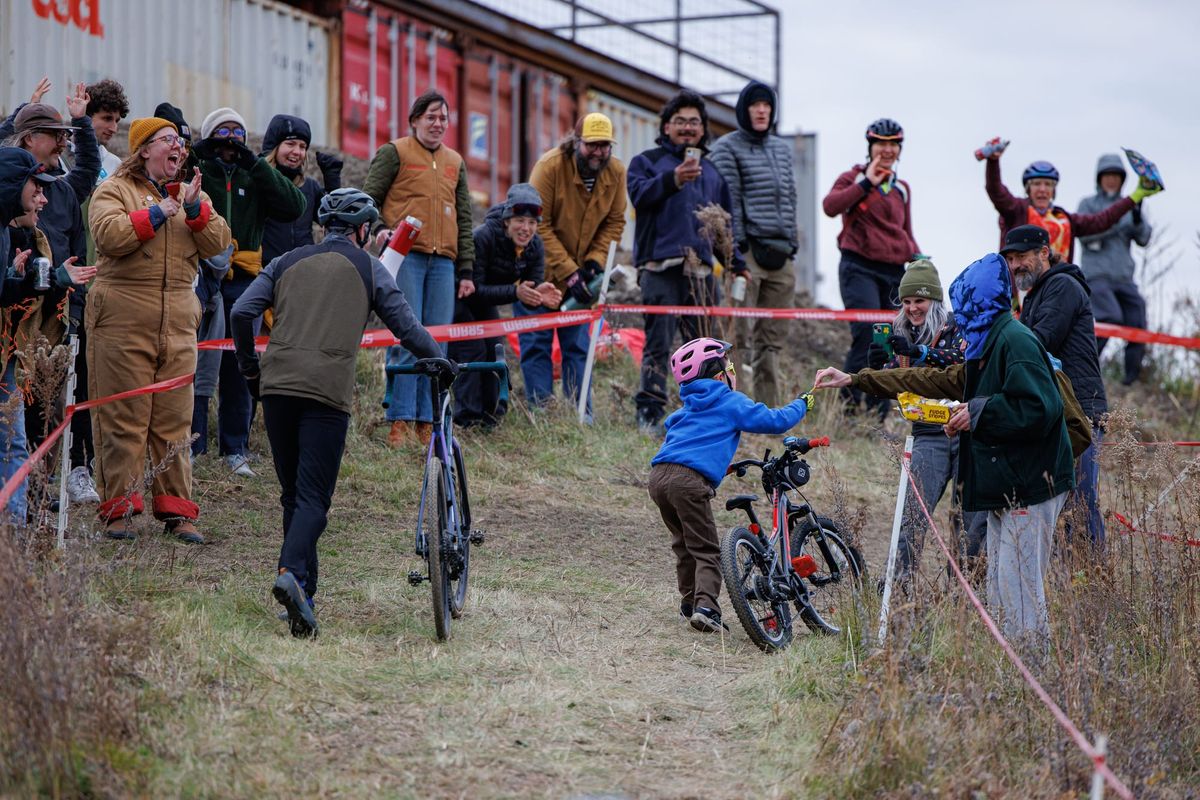 An adult and a child run up a hill with their bikes while spectators cheer and offer candy