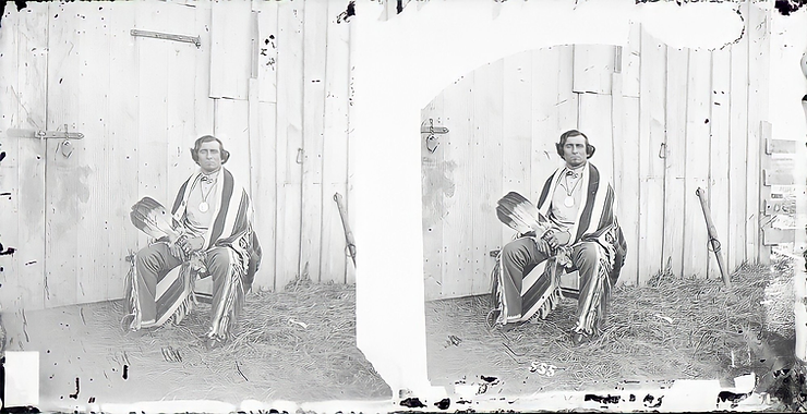 Baptiste Bayhylle seated in front of a wooden building, holding an eagle feather fan and wearing a blanket.