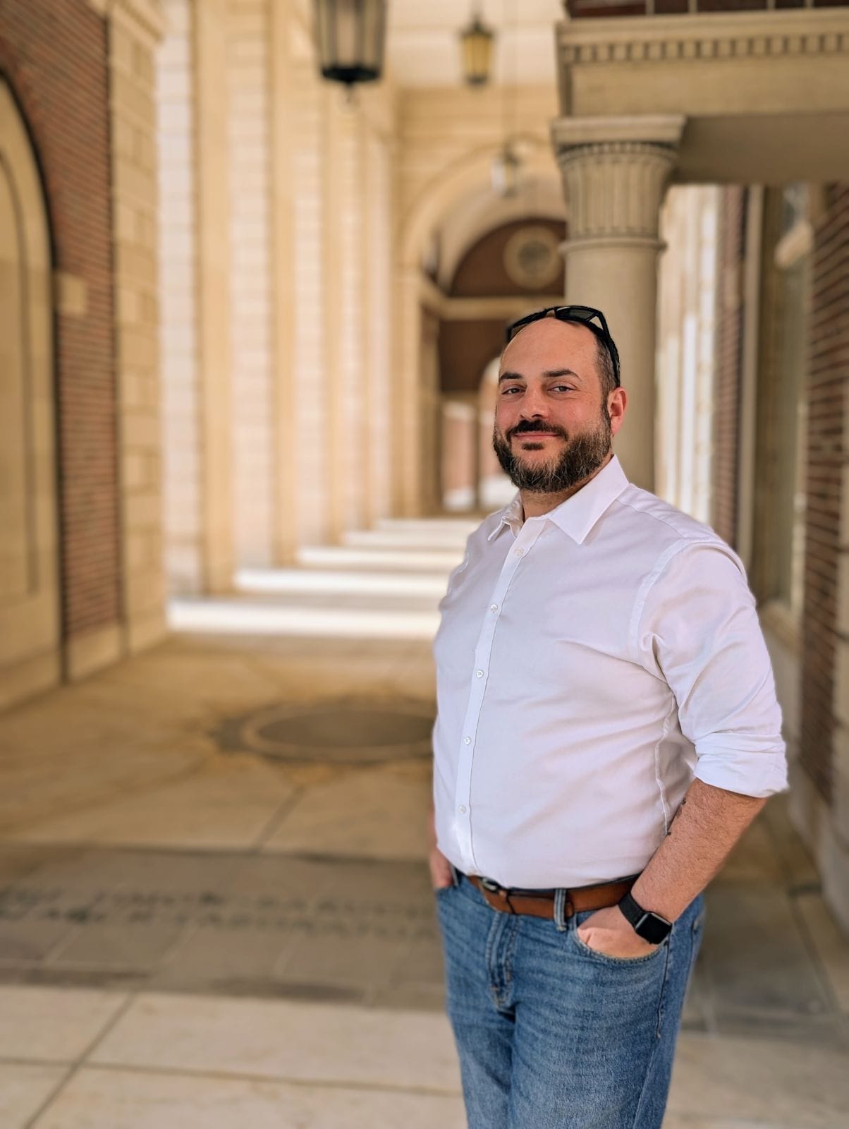 Photo of a while male presenting person, wearing a white button down oxford shirt, light blue denim jeans, a brown belt, and a pair of sunglasses propped on his forehead. He has dark brown/black hair and beard - the beard has a little salt and pepper in it - all close cropped. 