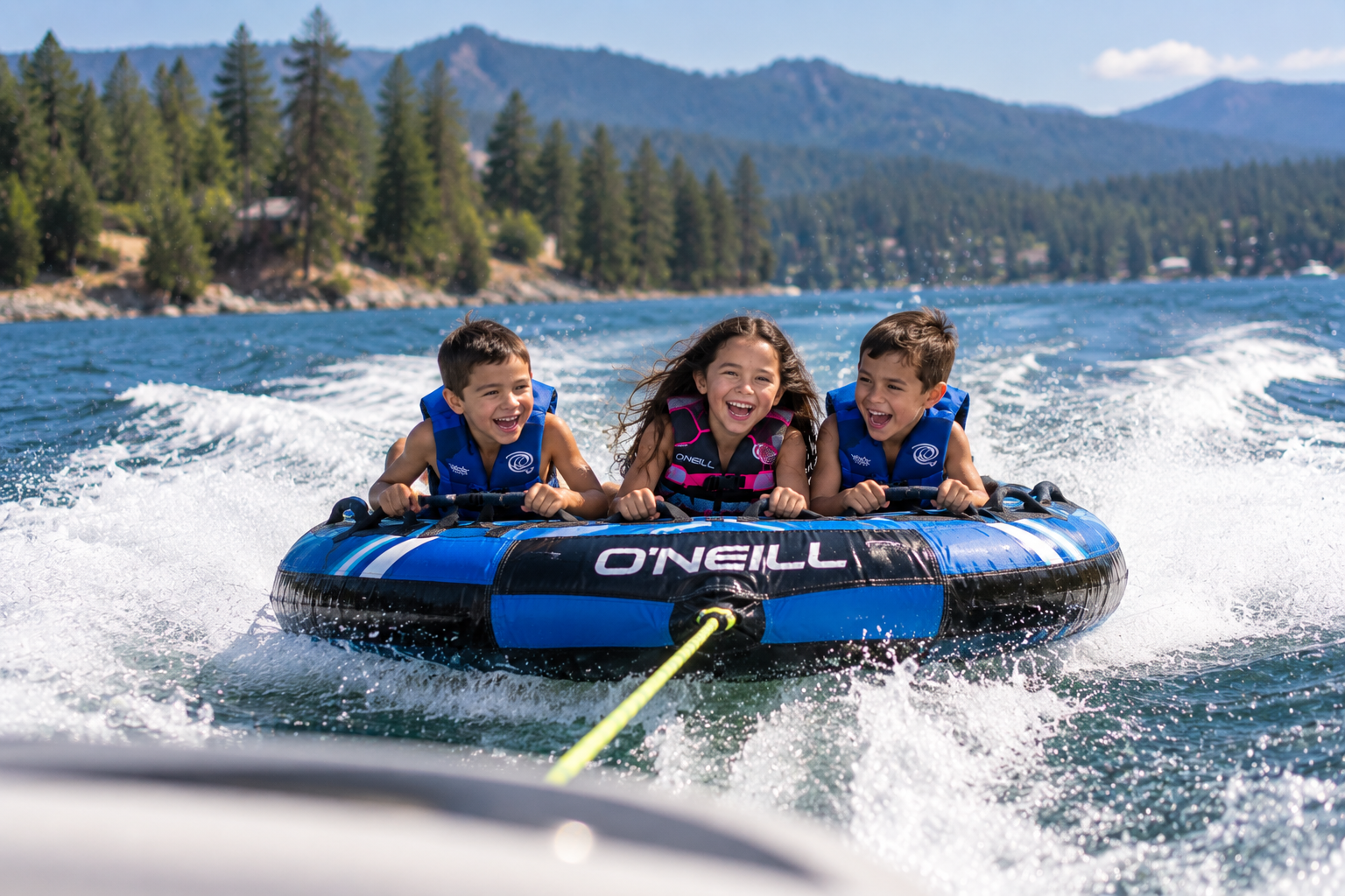 Three children wearing life jackets riding a towable tube behind a boat on a mountain lake.