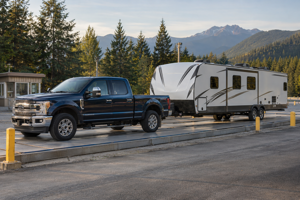 Pickup truck and travel trailer on an outdoor vehicle scale, with evergreen trees and mountains in the background.