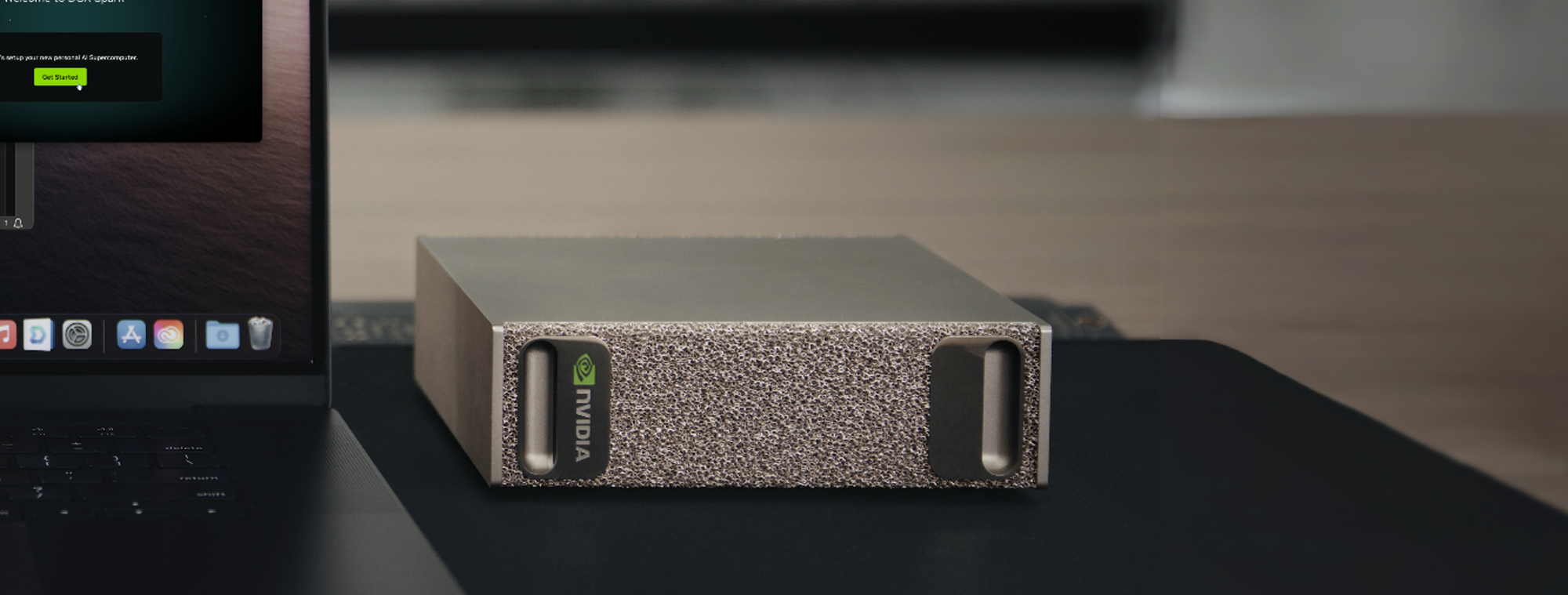 Close-up of an NVIDIA DGX Spark unit on a desk next to a laptop, showing its compact metallic design with textured front panel and NVIDIA branding.