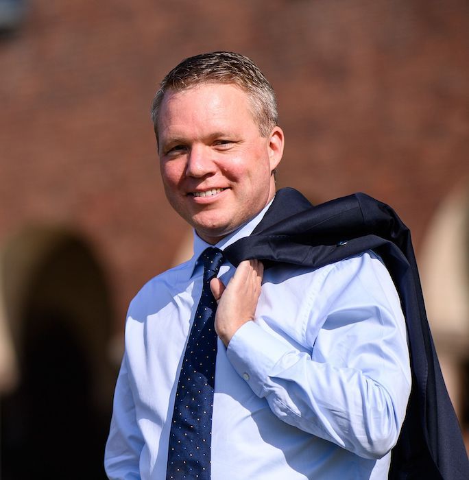 Headshot of Anders Larsson, in a blue shirt and blue polka dot tie, smiling with his suit jacket slung over his shoulder.