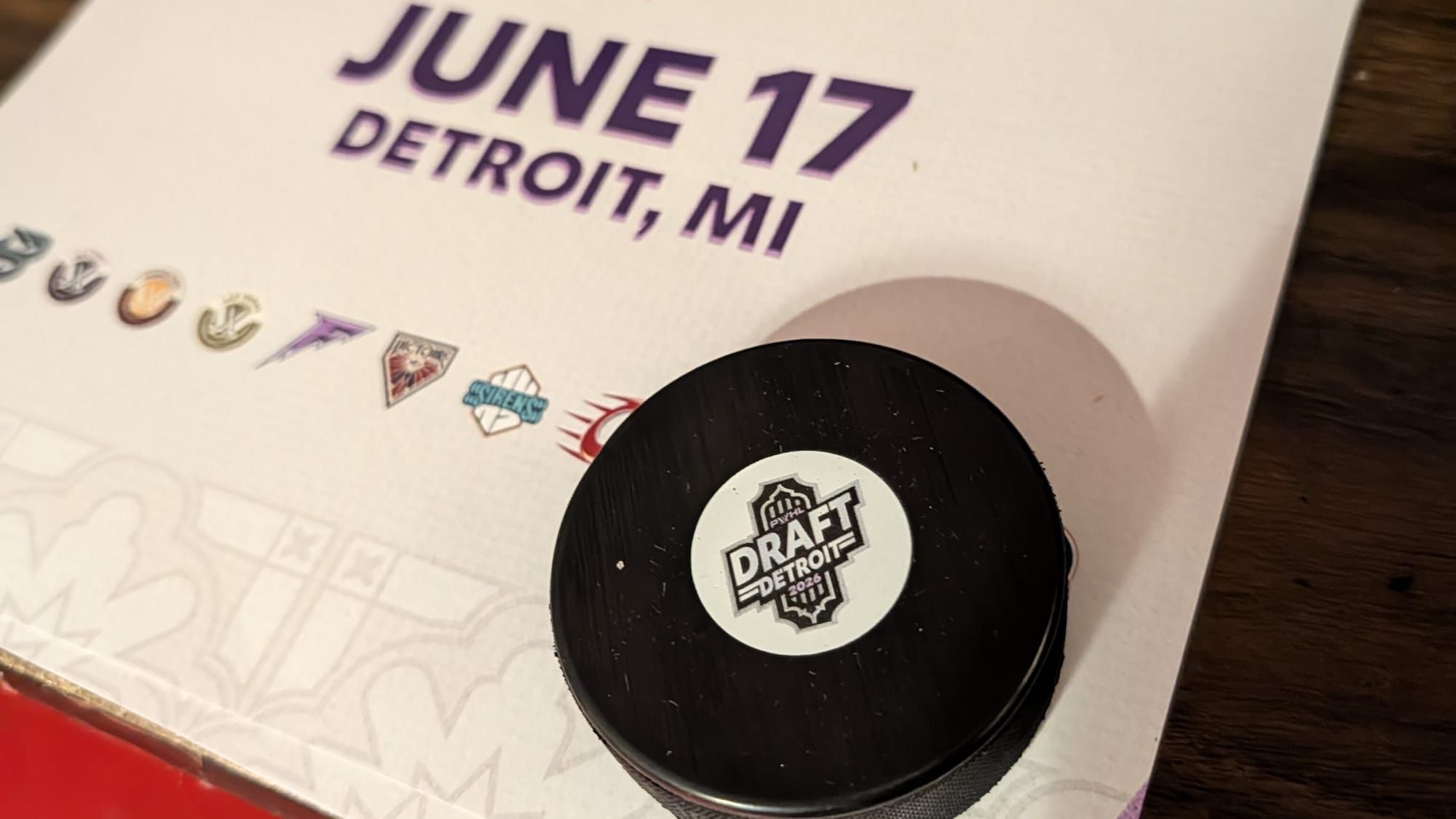 A hockey puck branded "PWHL Draft Detroit 2026" sits atop a white piece of paper with PWHL team logos and "JUNE 17 Detroit, MI" written in purple font.