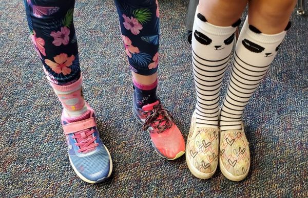 Two pairs of legs in shoes. On the left a person wears floral leggings, two socks of different lenghths and mismatched shoes. On the right a person wears slip-on shoes and panda knee socks.