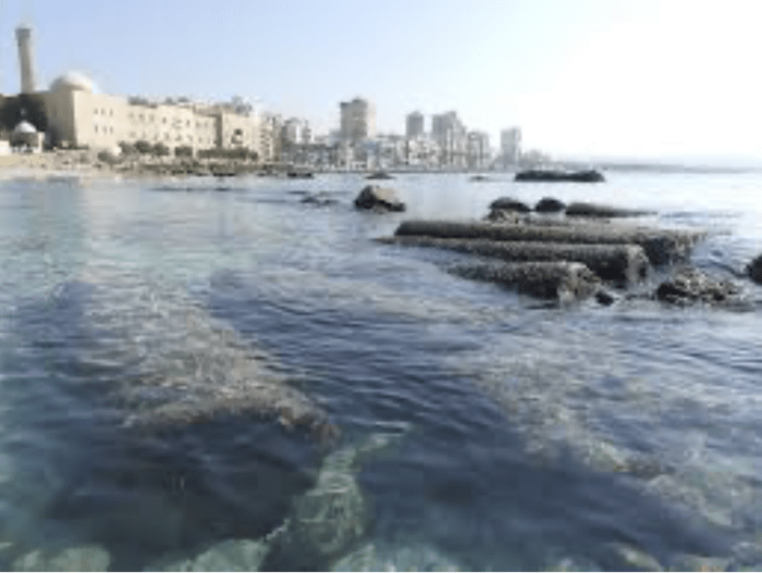 Bare rock of ancient columns out in the sea of the old city of Tyre with the skyline of a nearby modern city next to it in the background.
