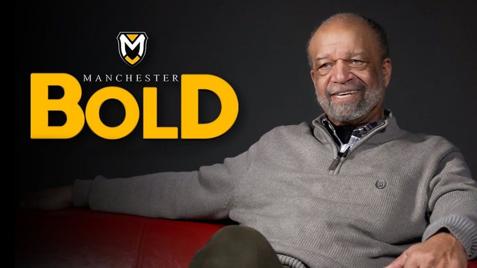 Myron Chenault, sitting with a smile in a gray sweater and olive pants.  The words "Manchester BOLD" are to his right.