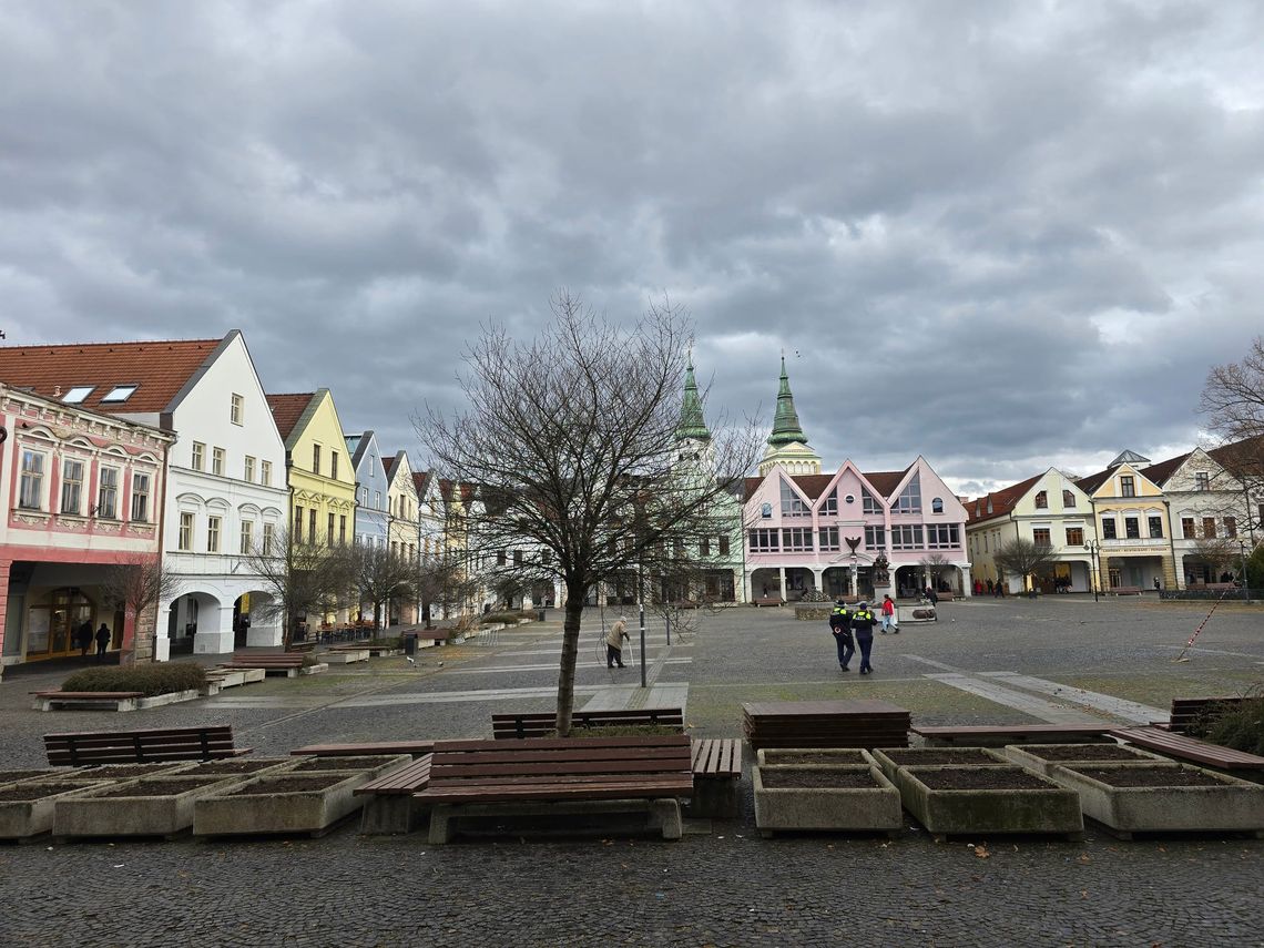 Town square with colorful buildings, a tree without leaves, church spires in background, and people walking.