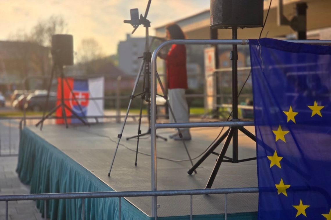Woman on stage with EU and Slovak flag on either side speaking into microphone